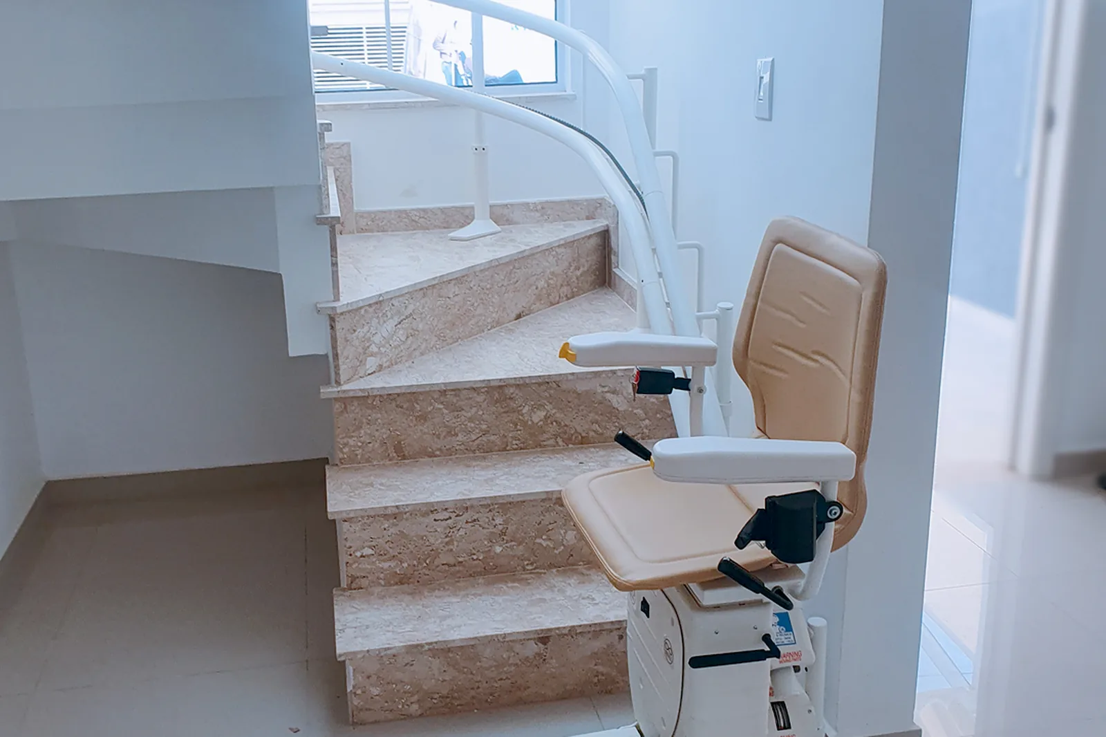 Treppenlift mit beigem Sitz am Antritt einer gewendelten Steintreppe, die Schiene ist auf den Stufen verschraubt